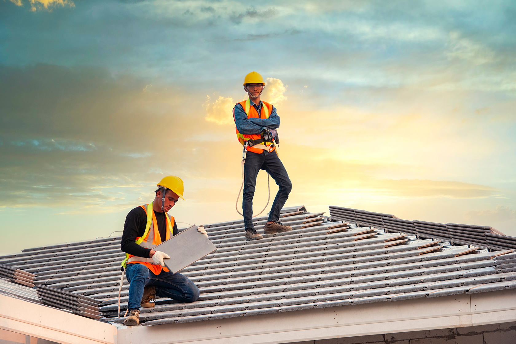 Two roofers working on a metal tile roof at sunset, wearing safety gear.